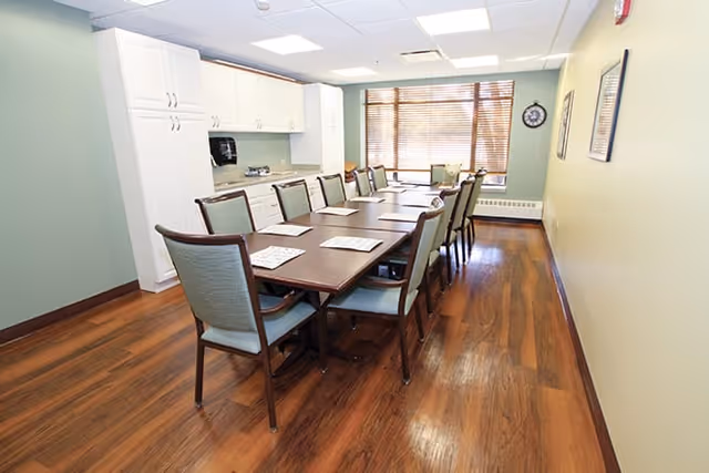 A long dining table with chairs arranged around it in a bright room with wooden flooring, white cabinets, and a large window with blinds letting in natural light.
