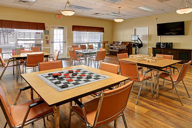 A well-lit common room with several tables and orange chairs arranged around them. One table has a checkers board set up, and another has a wooden block game. The room features large windows with blinds, a piano, a television mounted on the wall, and a door leading outside. The floor is wooden, and the ceiling has recessed lighting and hanging fixtures.
