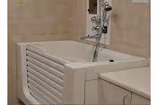 A walk-in bathtub with a side door and built-in faucet and showerhead, installed in a tiled bathroom corner next to a countertop with cabinets.