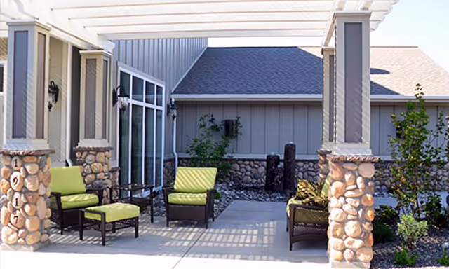 Outdoor patio area at Serenity Place featuring cushioned chairs and ottomans arranged under a pergola with stone pillars, adjacent to a building with stone and siding exterior walls and some greenery.