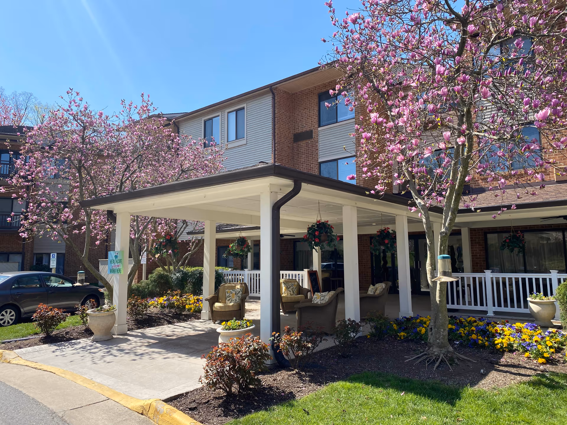 Outdoor covered seating area at the entrance of a senior living facility with blooming pink trees and colorful flower beds surrounding the space. The building has multiple windows and a brick and siding exterior. Several chairs with cushions are arranged under the covered area.