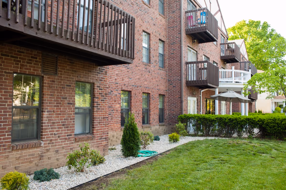 Brick apartment building with wooden balconies, landscaped shrub beds, and a grassy lawn.