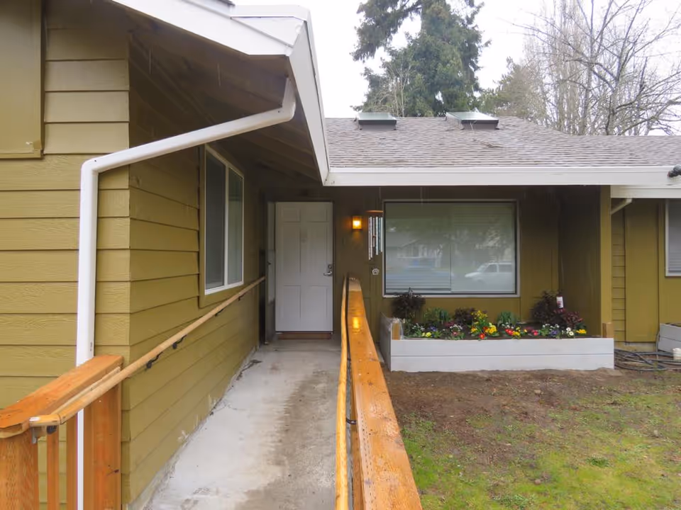 Exterior view of the entrance to Rumaan Adult Family Home showing a covered walkway with wooden handrails leading to a white door. There is a large window with closed blinds and a flower bed with colorful flowers beneath it. The building is painted olive green and there are trees visible in the background.