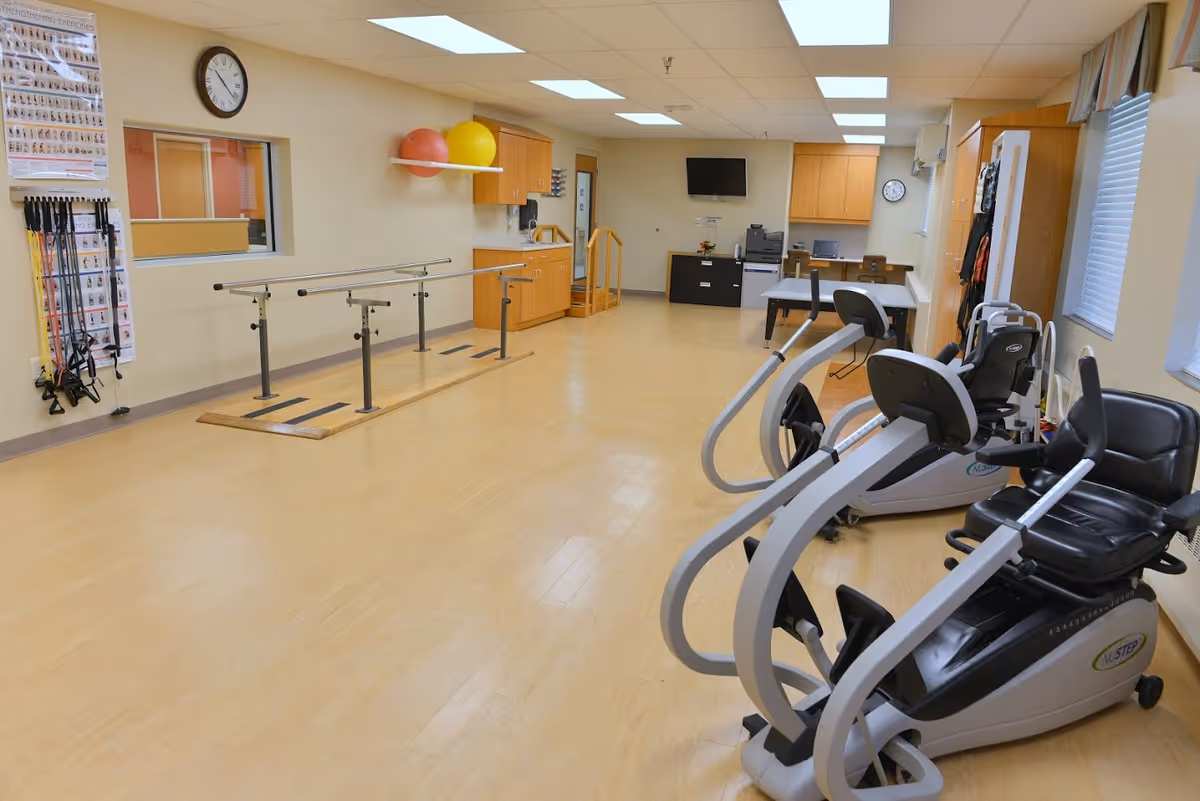 A spacious rehabilitation or physical therapy room with wooden flooring, parallel bars for walking exercises, exercise balls on a wall shelf, resistance bands hanging on the wall, two recumbent exercise bikes, a desk with a computer and printer, and a wall-mounted TV. The room is well-lit with ceiling lights and has windows with blinds.