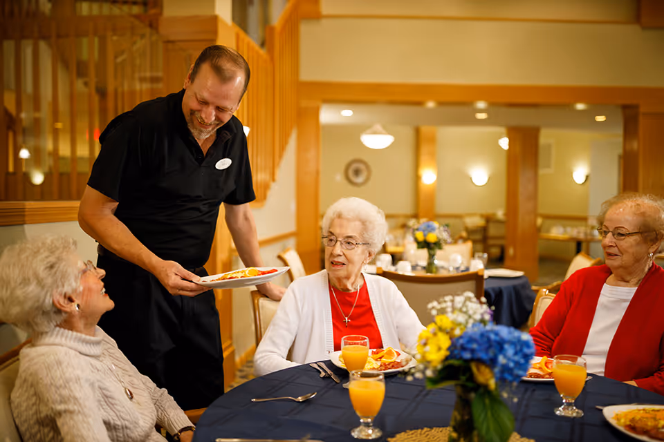 A male server in a black uniform smiling and serving breakfast to three elderly women seated at a round table with a dark blue tablecloth in a dining room. The table has glasses of orange juice, plates of food, and a floral centerpiece with blue and yellow flowers. The background shows a warmly lit dining area with wooden accents and more tables.