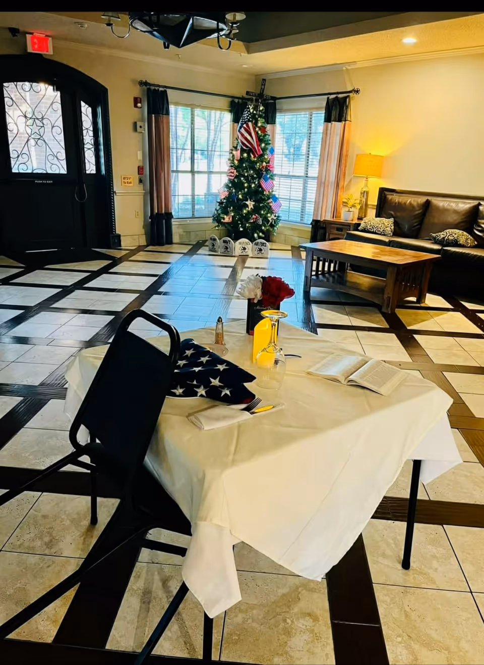 A decorated indoor space with a table covered by a white tablecloth, set with a folded American flag, a glass, a napkin, and an open book. In the background, there is a Christmas tree adorned with American flags and ornaments, a black leather couch with patterned pillows, a wooden coffee table, and large windows with curtains letting in natural light. The floor has a tiled pattern with dark and light tiles, and a black door with decorative glass is visible on the left.