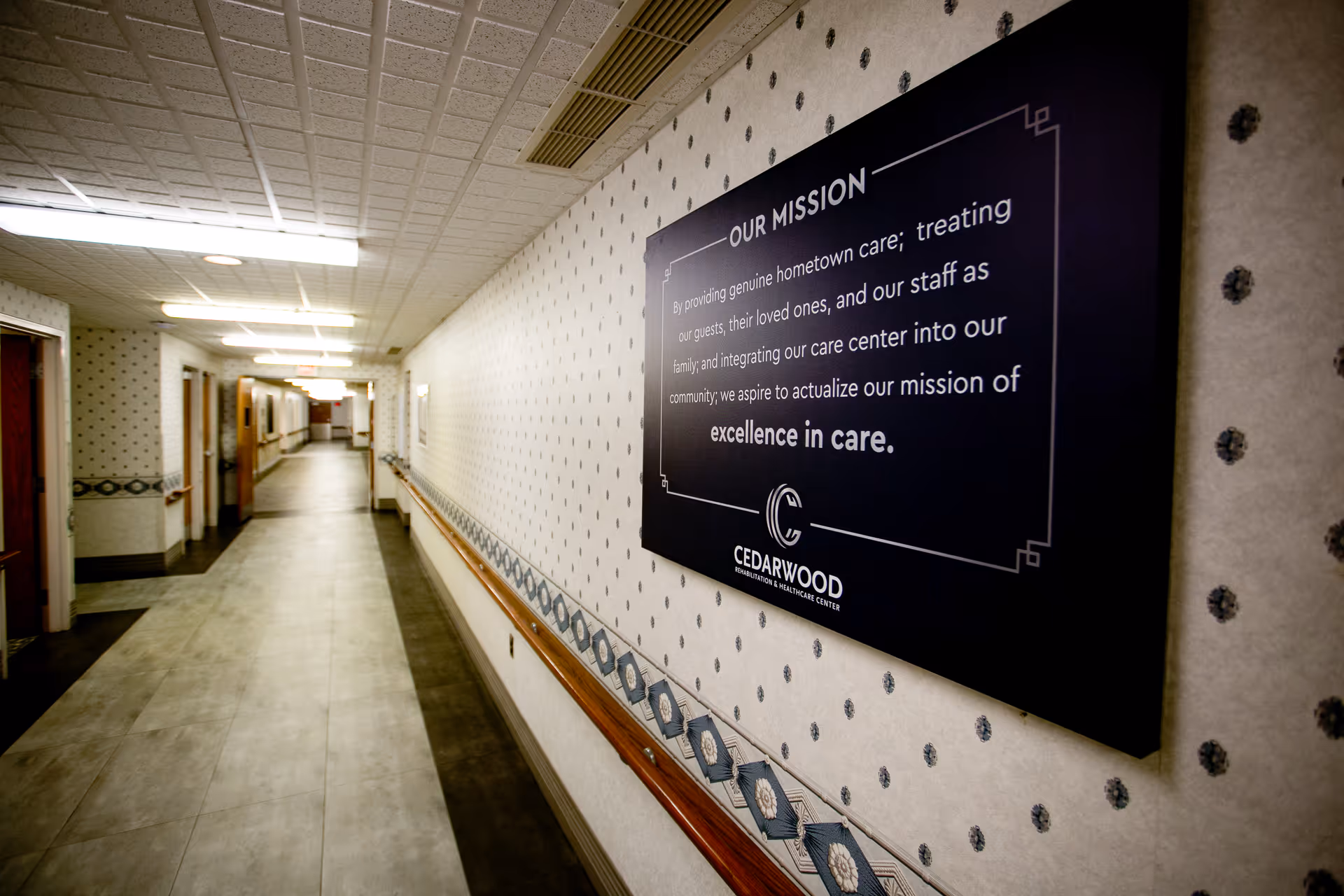 A long, empty hallway in Cedarwood Rehabilitation and Healthcare Center with patterned wallpaper, handrails along the walls, and a large sign displaying the facility's mission statement.