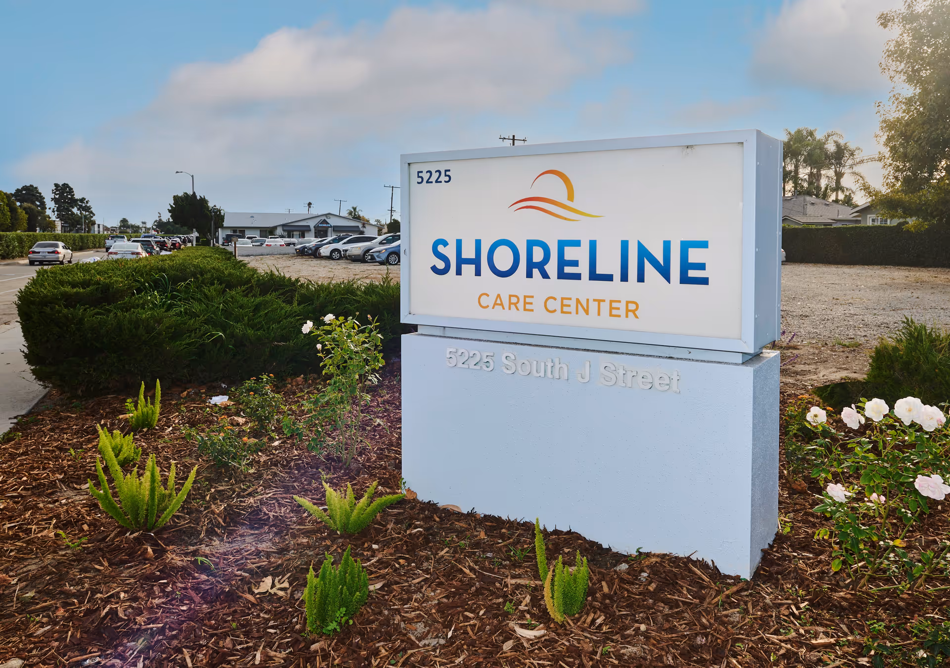Outdoor view of the Shoreline Care Center sign with the address 5225 South J Street. The sign is surrounded by landscaped plants and bushes, with a parking lot and building visible in the background under a partly cloudy sky.