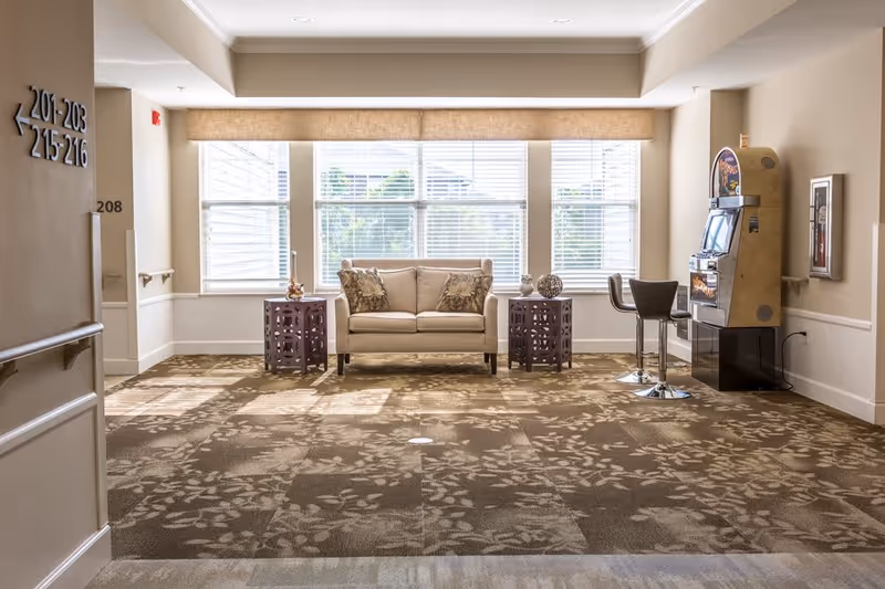 Bright senior living lounge with a sofa and side tables in front of large windows and a gaming machine to the right.