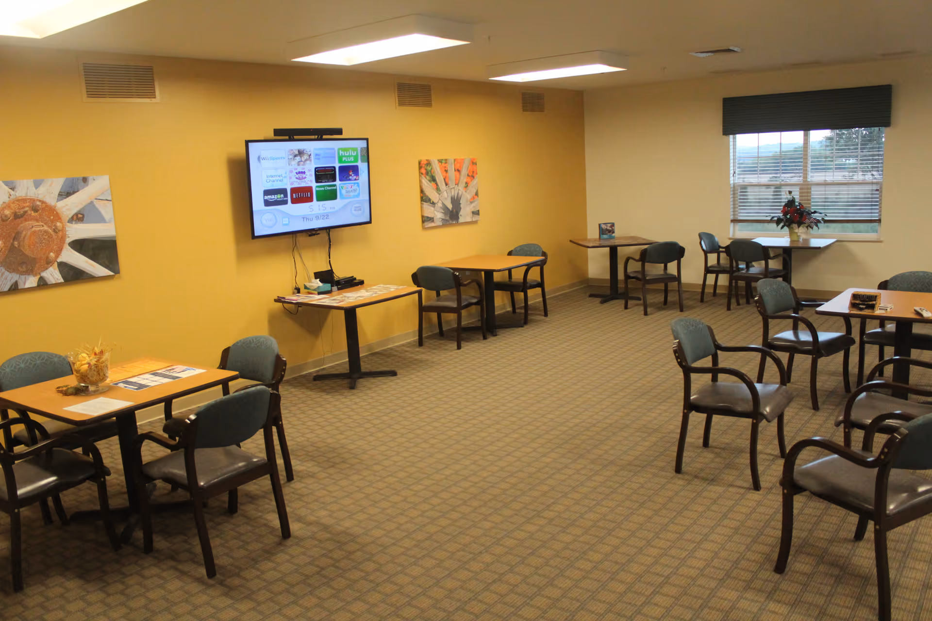 Communal activity/dining room with multiple tables and chairs, a wall-mounted TV, and yellow walls.