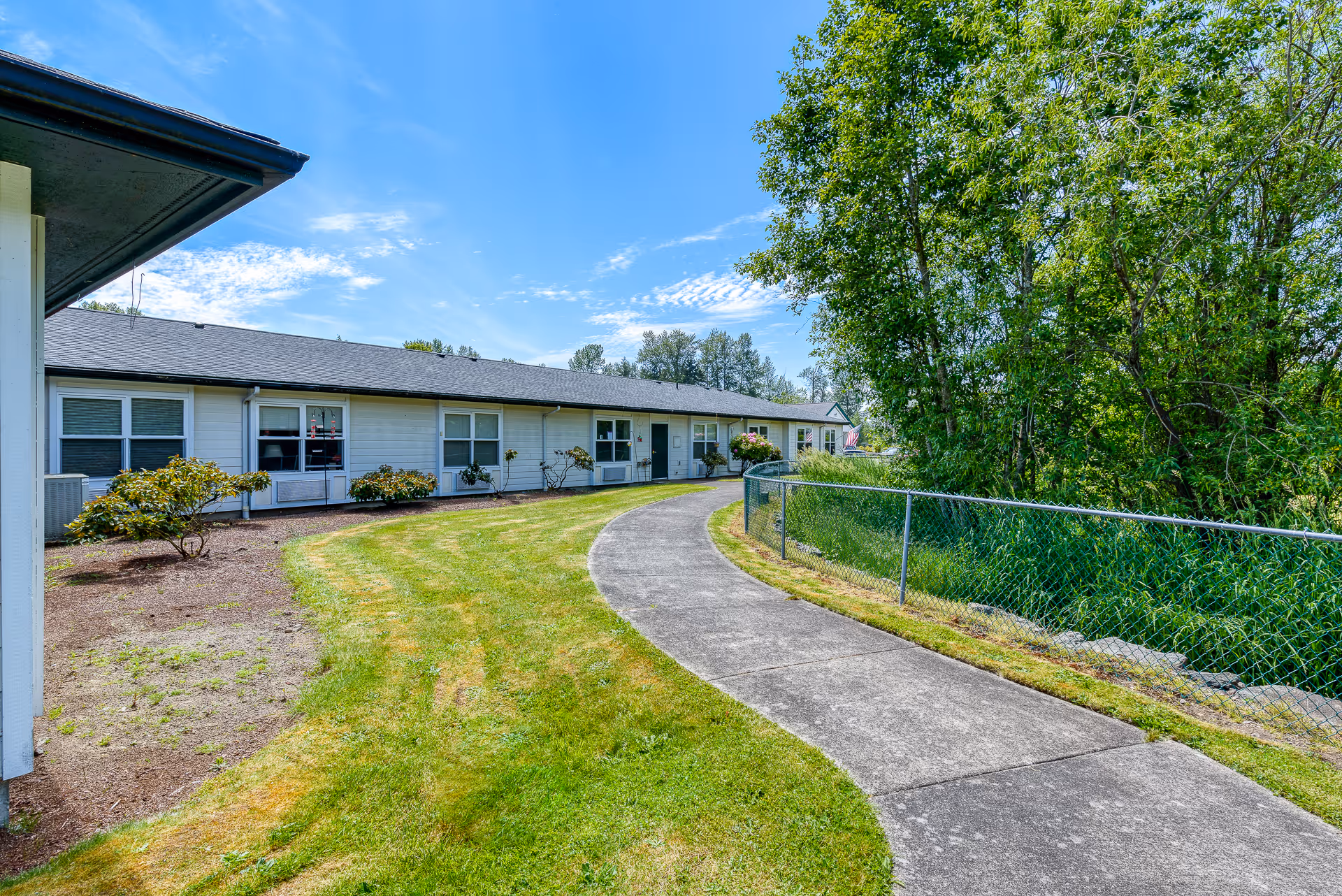 Curved concrete pathway bordered by green grass and a chain-link fence on the right, leading to a single-story building with white siding and multiple windows under a clear blue sky.