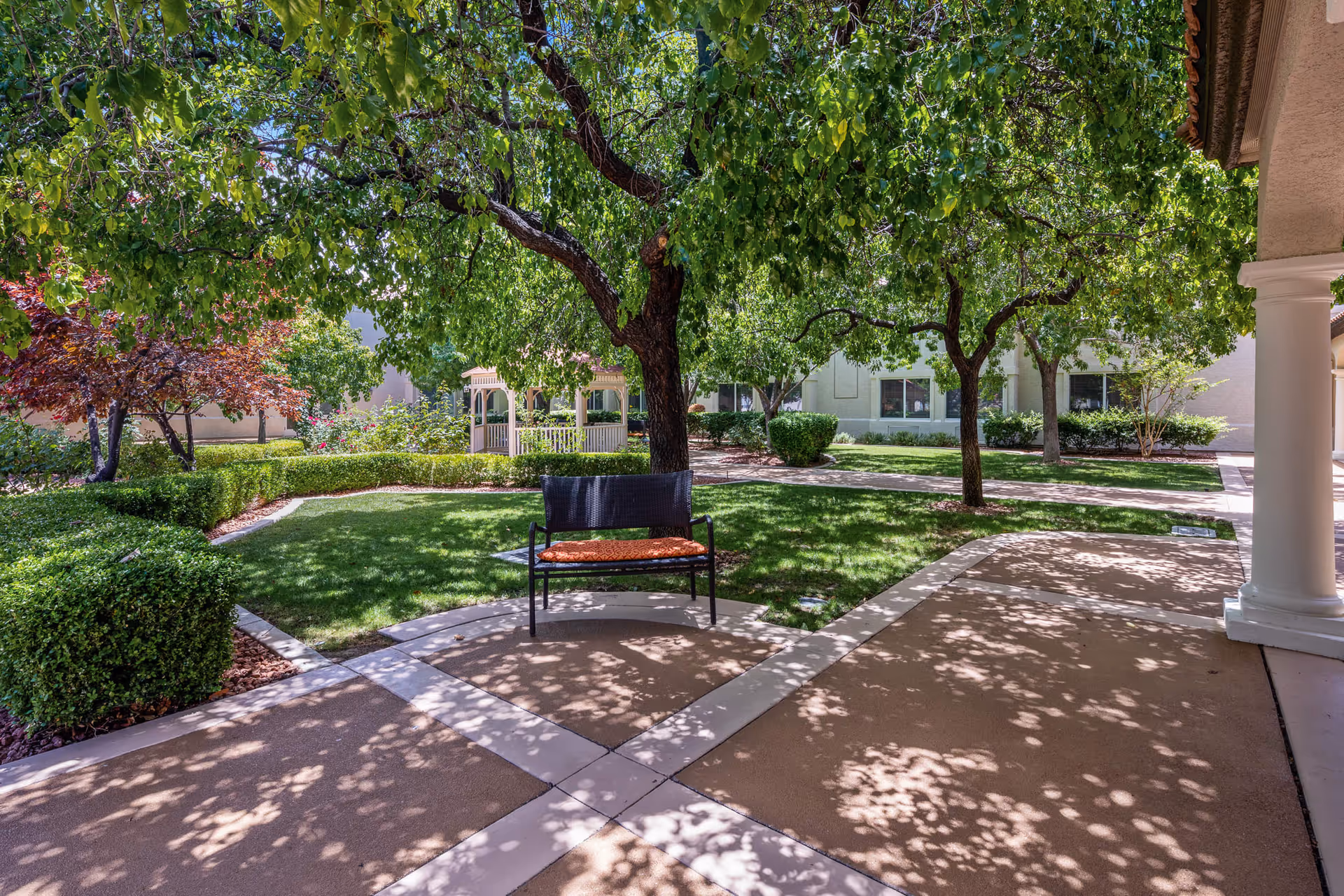 A peaceful outdoor garden area with a bench under the shade of leafy green trees. The garden features well-maintained grass, bushes, and a gazebo in the background. There are paved walkways and a building with windows visible behind the garden.