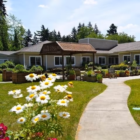 Outdoor garden area with a curved concrete pathway leading to a pergola structure surrounded by raised flower beds and chairs. The background shows a single-story building with windows and a roof, with trees and a partly cloudy sky behind it.