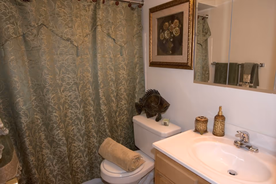 Bathroom with a toilet, a rolled beige towel on the toilet seat, a decorative fish sculpture on the toilet tank, a sink with a faucet, soap dispenser, and a small container on the countertop. There is a framed floral painting on the wall and a green patterned shower curtain in the background.