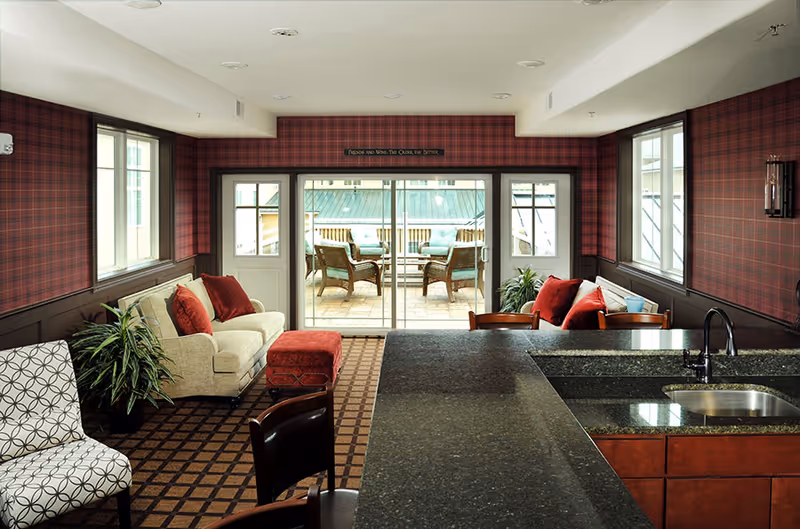 Interior view of a senior living facility lounge area with plaid wallpaper and carpet. The room features two beige sofas with red cushions, a patterned armchair, a red ottoman, and a granite countertop with a sink in the foreground. Large sliding glass doors lead to an outdoor patio with wicker chairs and a table.