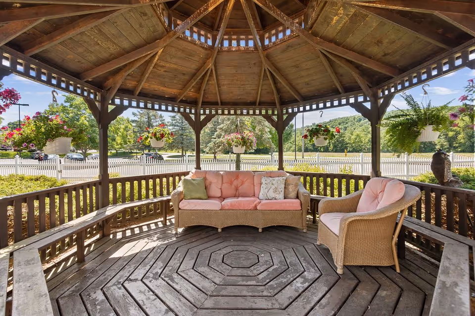 A wooden gazebo with a hexagonal roof and wooden floor, furnished with a wicker sofa and chair with pink cushions. Hanging flower pots with colorful flowers are suspended around the gazebo, and a white picket fence and green trees are visible in the background under a clear blue sky.