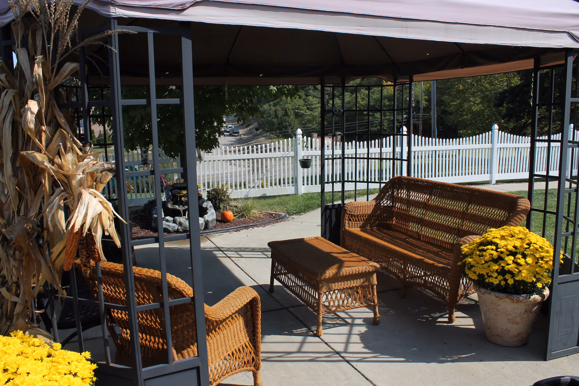 Outdoor seating area under a canopy with wicker furniture including a loveseat, chair, and coffee table. There are yellow flowers in a pot and dried corn stalks decorating the area. A white picket fence and greenery are visible in the background.