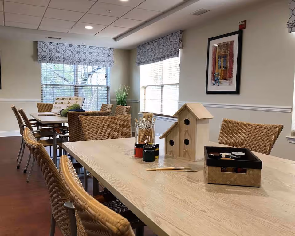 Bright interior dining/activity room with wicker chairs and tables set up for crafts including wooden birdhouses, paint and brushes by large windows.