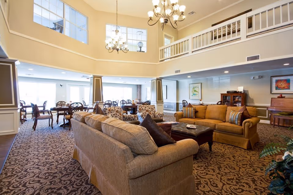 Spacious senior living facility common area with two large beige sofas, patterned armchairs, a dark ottoman, and a dining area with multiple tables and chairs. The room features high ceilings with large windows, two chandeliers, and decorative wall art.