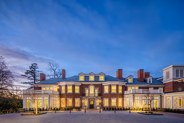 Illuminated red-brick senior living building front at dusk with symmetrical windows, porches, and a circular driveway.
