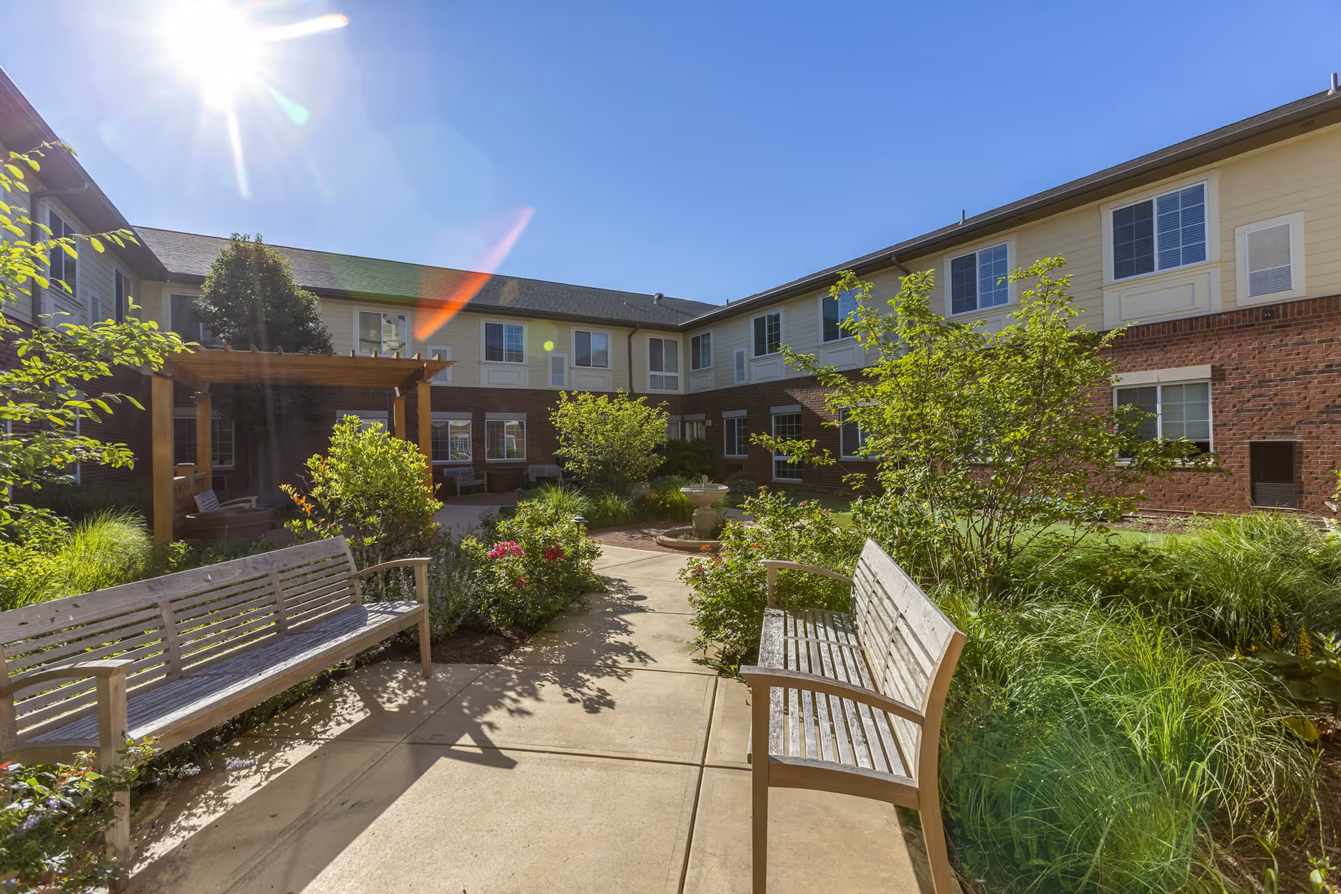 Sunny outdoor courtyard at Independence Village of Zionsville West featuring two wooden benches, a paved walkway, lush green plants, flowers, and a wooden pergola attached to the building.
