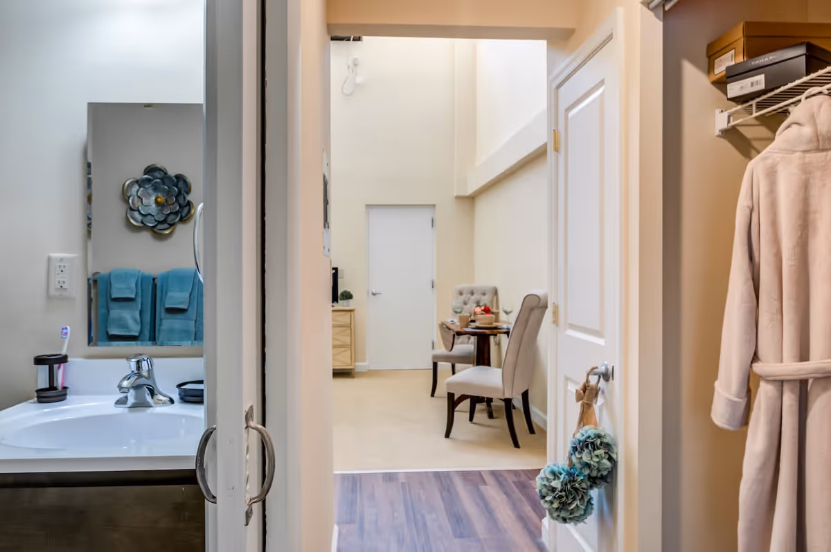View down a hallway in a senior living facility showing a bathroom sink with a mirror and blue towels on the left, a small dining area with two upholstered chairs and a table in the background, and a closet on the right with a hanging beige robe and storage boxes on a shelf.