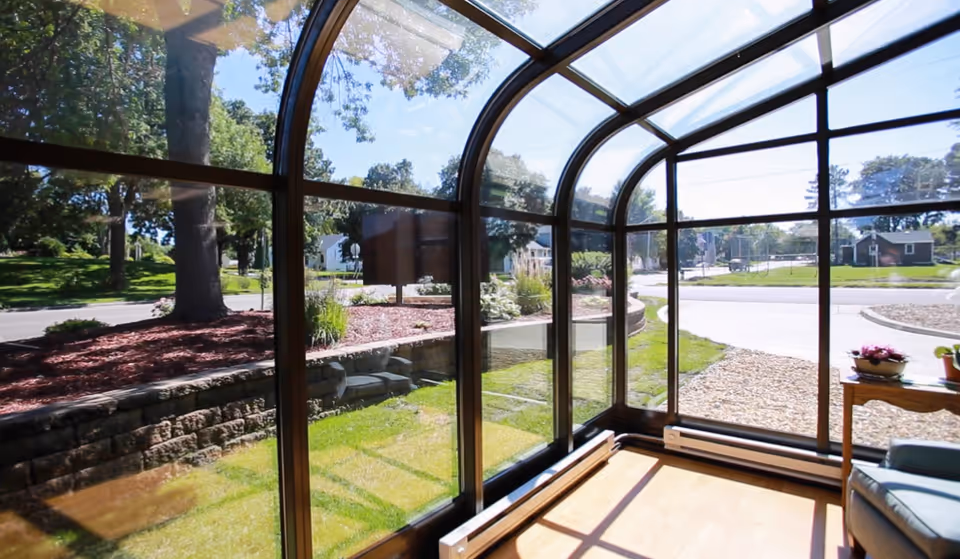 A sunroom with large curved glass windows providing a clear view of a landscaped outdoor area with grass, trees, and a stone retaining wall. The room has a wooden floor, a small table with potted plants, and a cushioned chair.