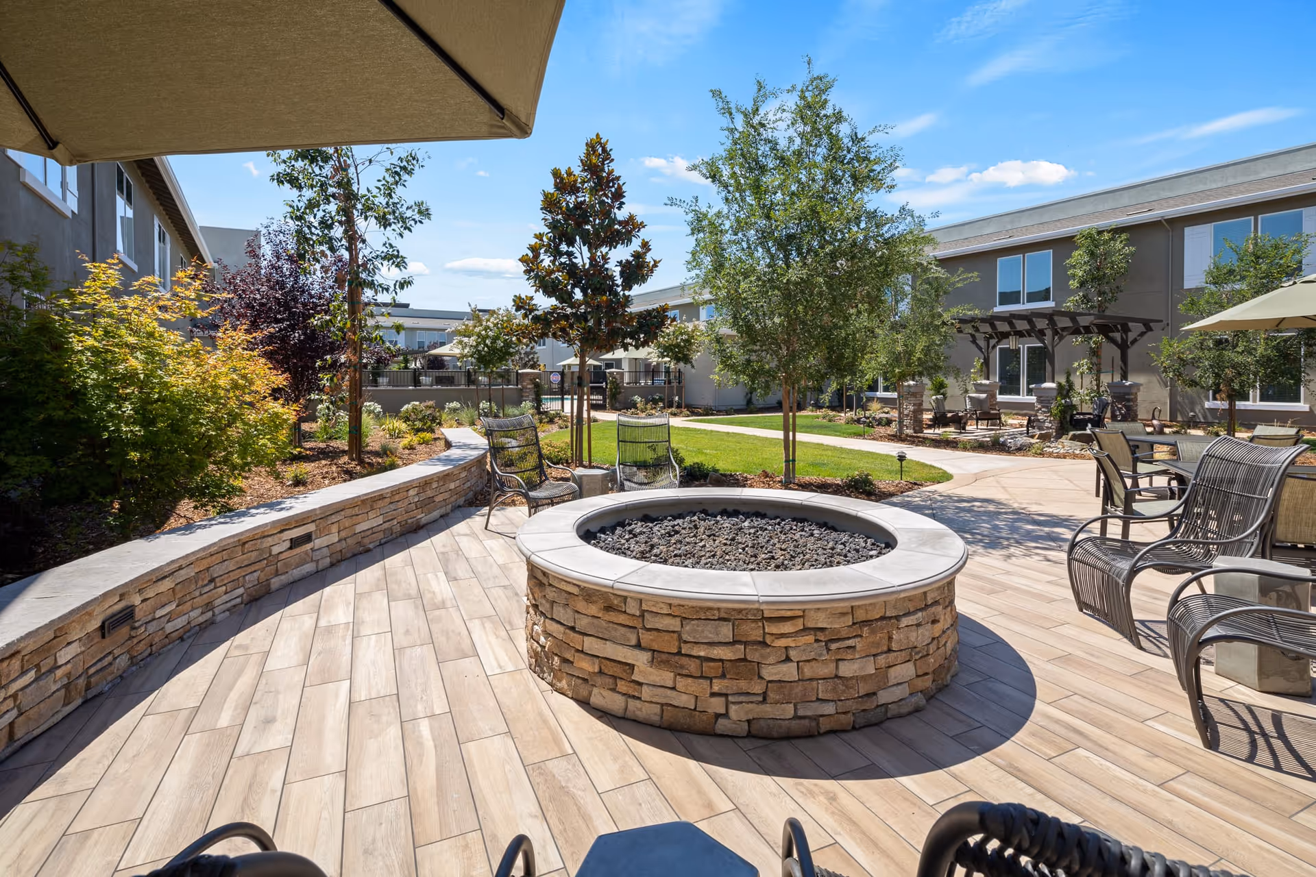 Outdoor patio area at Oakmont of Lodi featuring a circular stone fire pit surrounded by metal chairs, a stone bench, trees, shrubs, and umbrellas under a clear blue sky.