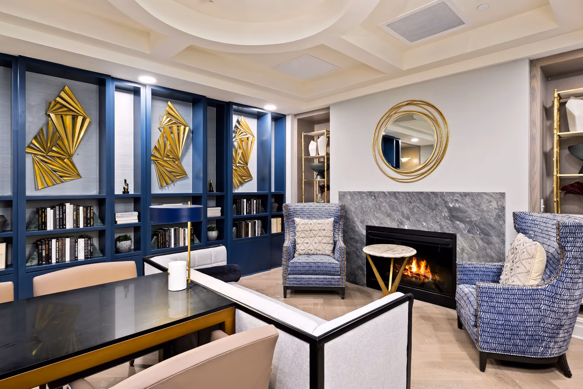 A cozy living room area with a modern fireplace surrounded by gray stone. Two blue patterned armchairs with cream-colored pillows face the fireplace, with a small round marble side table between them. A large round gold-framed mirror hangs above the fireplace. To the left, there is a black table with beige chairs and a white lamp. The back wall features built-in blue shelves filled with books and decorative items, accented by three gold geometric wall sculptures.