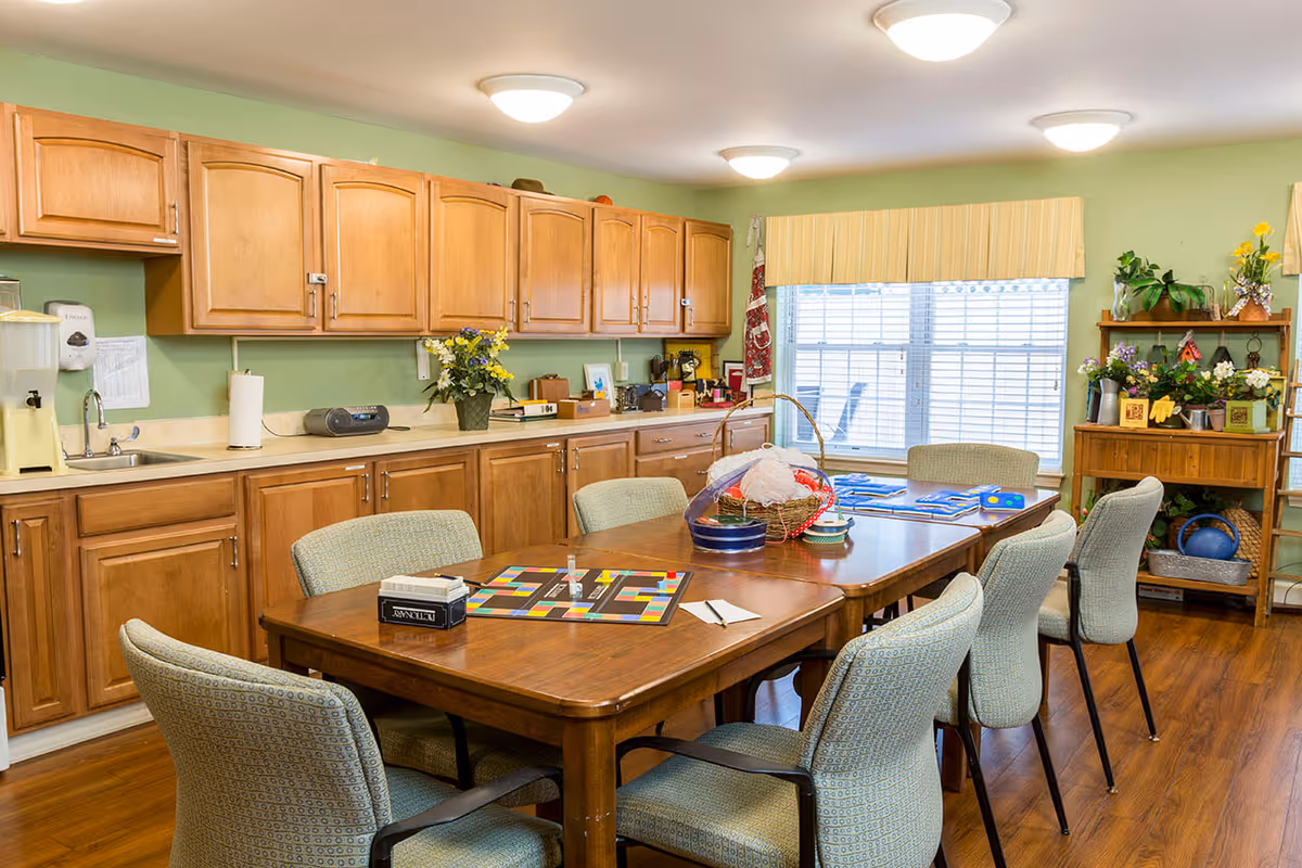 Communal dining/activity room with a wooden table surrounded by upholstered chairs, a kitchenette with wooden cabinets, and a window with blinds.