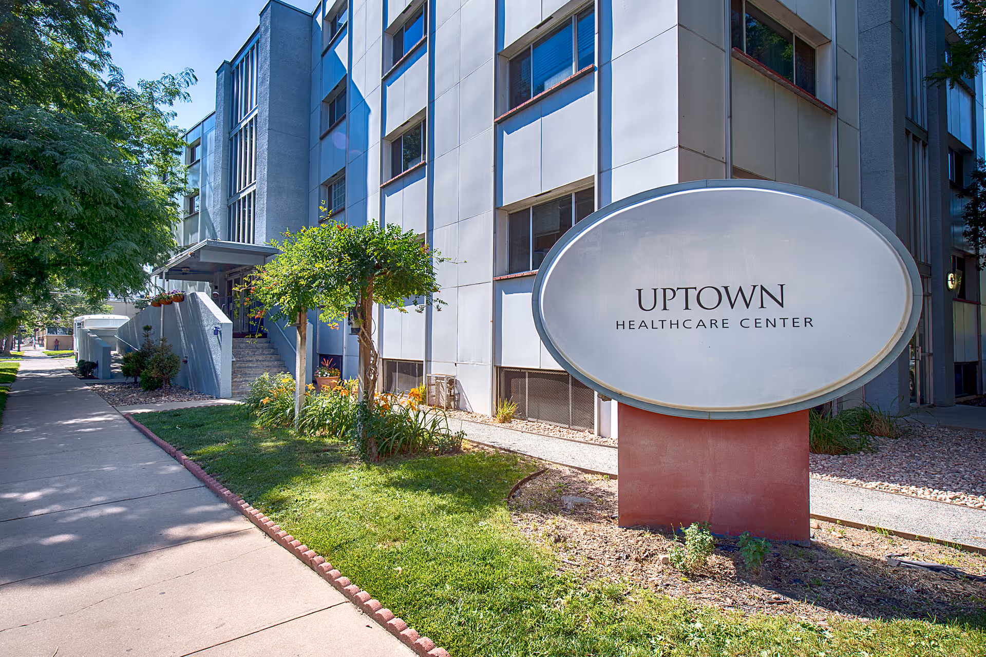 Exterior view of Uptown Healthcare Center building with a large oval sign displaying the facility name. The building is multi-story with windows and surrounded by greenery including trees and grass along a sidewalk.