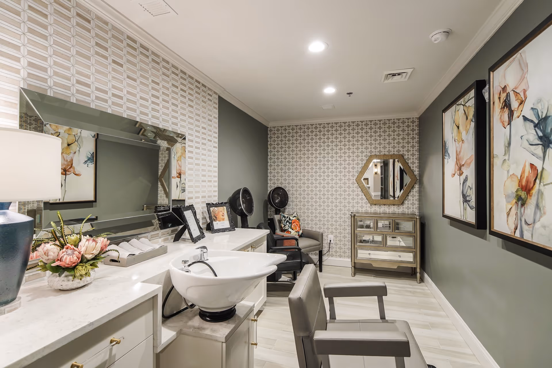 Interior view of a salon area in a senior living facility with a white sink and salon chair in the foreground, a large mirror on the left wall, two hair dryers, a small cabinet with a hexagonal mirror above it, and floral artwork on the right wall. The room has patterned wallpaper and soft lighting.