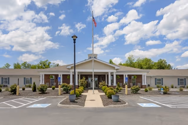 Front exterior view of a single-story senior living facility building with a central entrance featuring a covered porch supported by white columns. There is a flagpole with an American flag in front of the entrance, surrounded by landscaped bushes and plants. Handicap parking spaces are visible on either side of the walkway leading to the entrance. The sky is partly cloudy with blue patches.