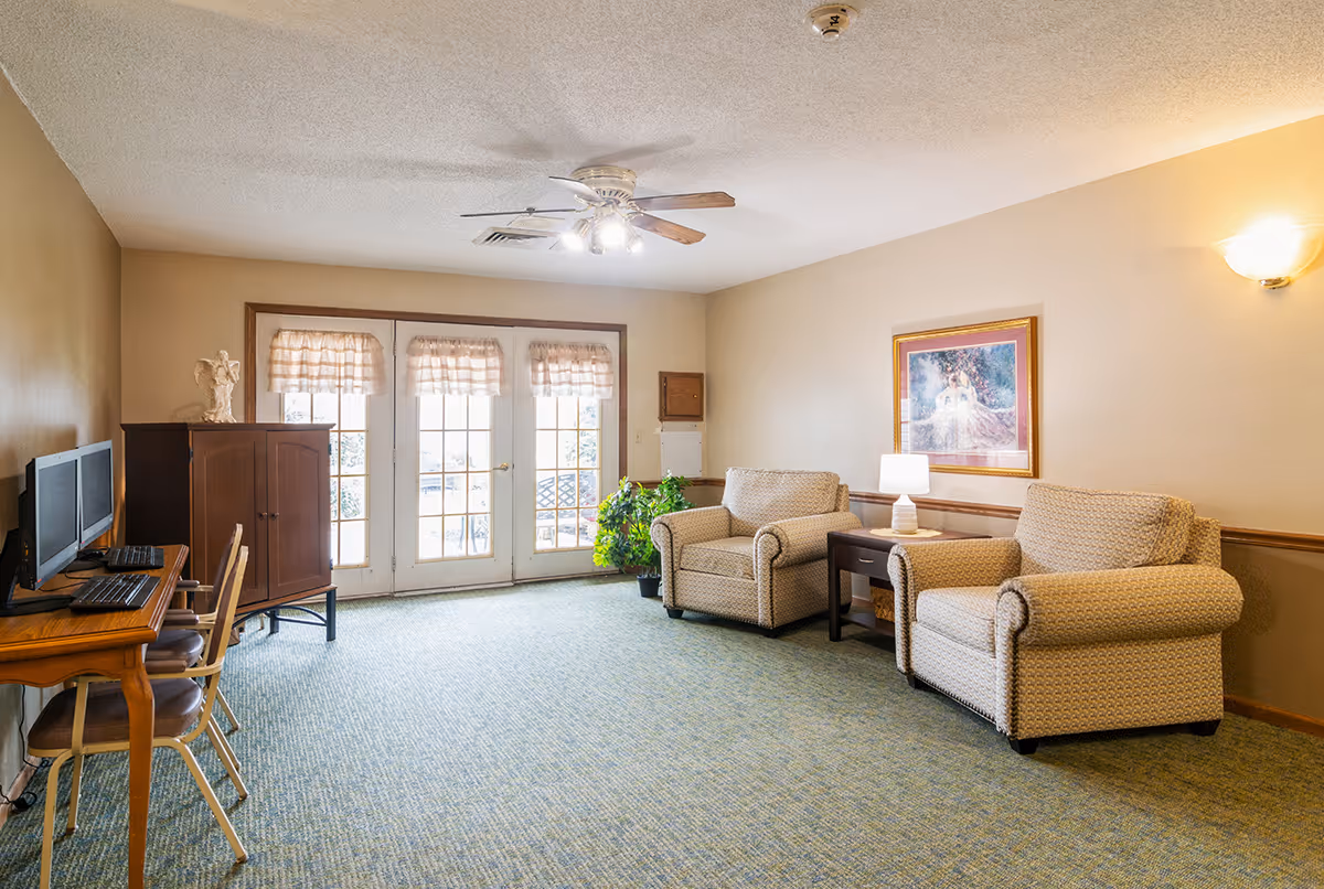 A cozy living room area with two beige upholstered armchairs separated by a small wooden side table with a white lamp. A framed painting hangs on the wall above the chairs. To the left, there is a wooden desk with two computer monitors and chairs. French doors with sheer curtains allow natural light into the room. The floor is carpeted in a muted green pattern, and a ceiling fan with lights is mounted on the ceiling.