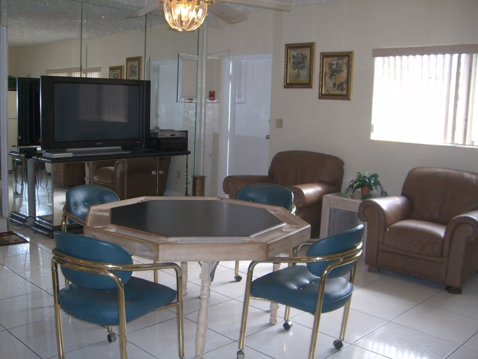 Interior living room with an octagonal game table surrounded by teal chairs, a television on a stand, and leather armchairs by a window.