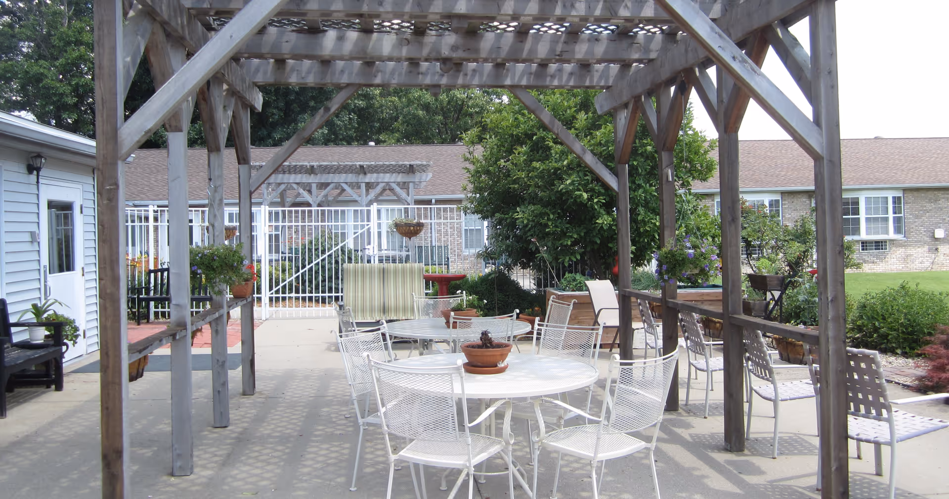 Outdoor courtyard with a wooden pergola, white metal patio tables and chairs, potted plants, and a low building in the background.