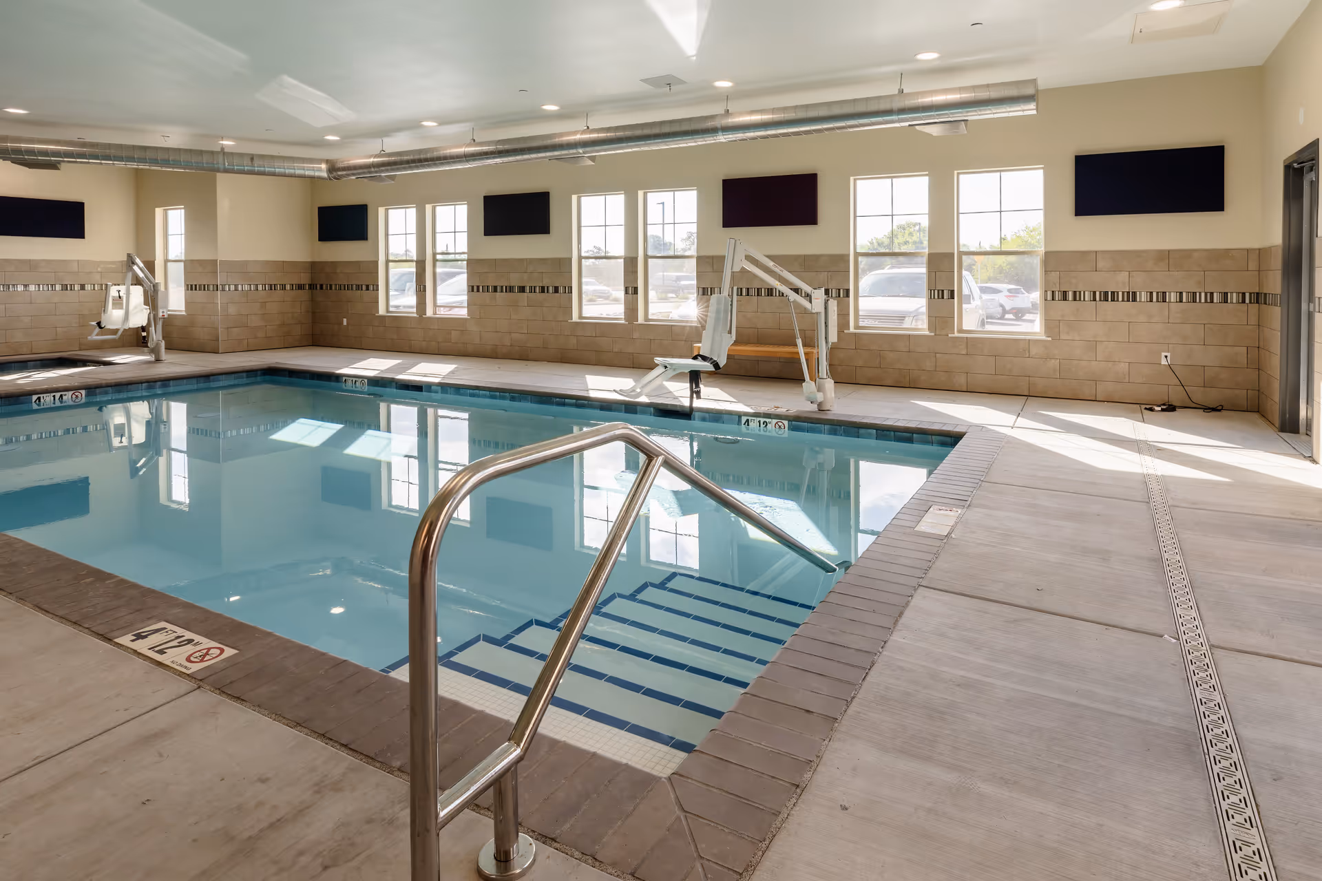 Indoor swimming pool area with clear blue water, metal handrails, tiled walls, and large windows letting in natural light. There are two pool lifts for accessibility and several black wall-mounted panels above the windows.