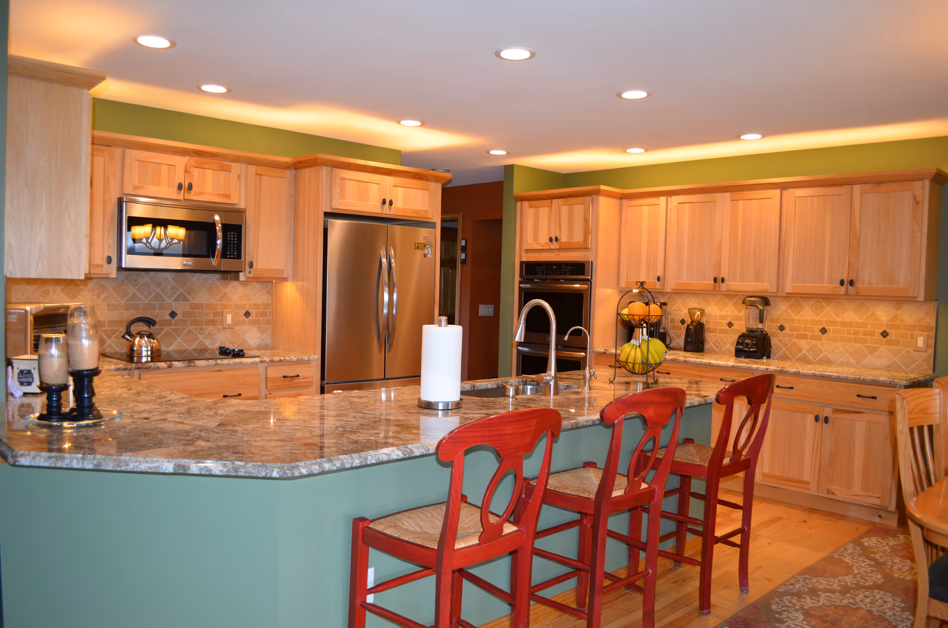Spacious kitchen with a green island and granite countertop, three red bar stools, wooden cabinets, and stainless steel appliances.