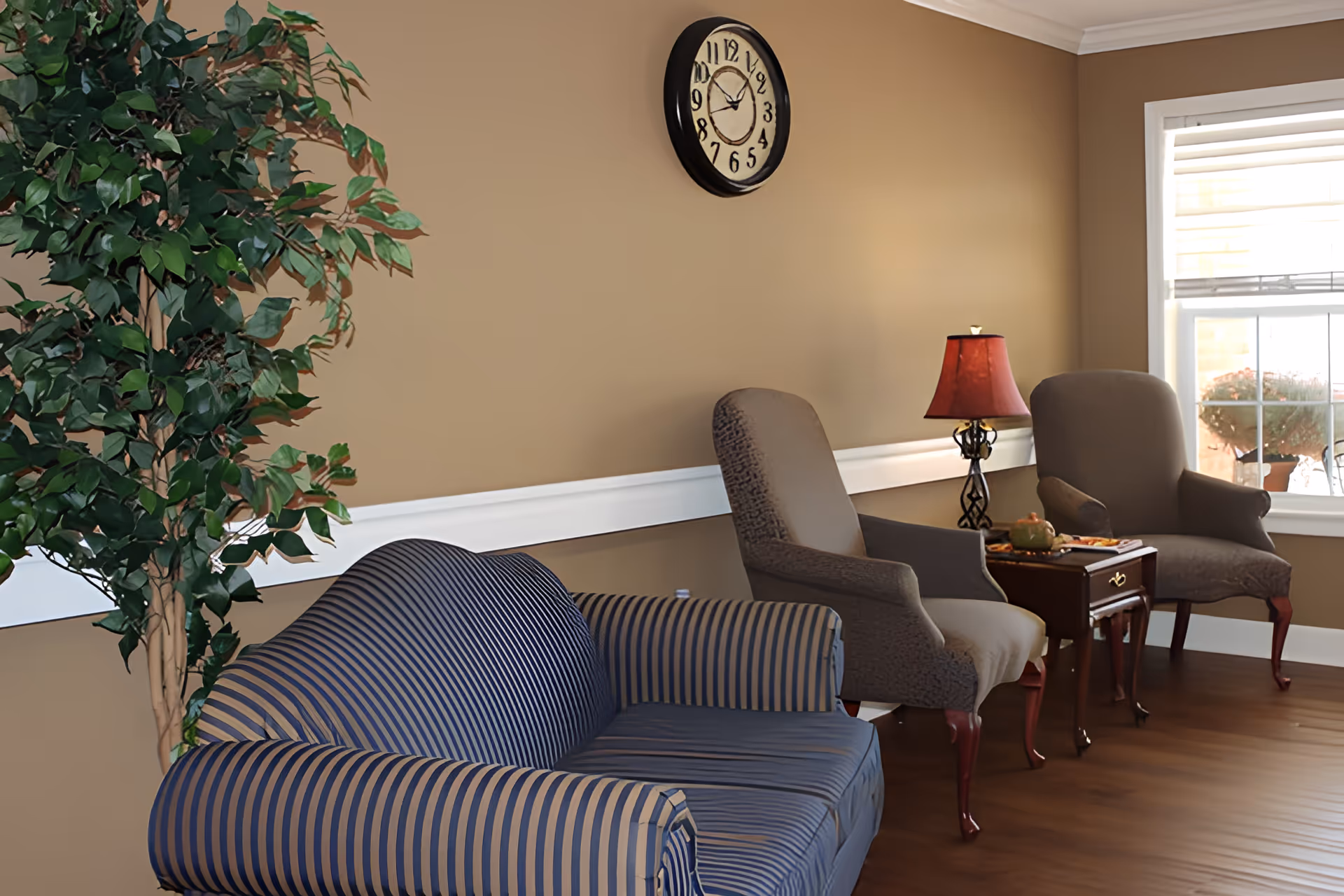 A cozy sitting area with a striped blue loveseat, two upholstered armchairs, a small wooden side table with a red lamp, and a large leafy plant against a beige wall with a round clock. A window with white trim lets in natural light.