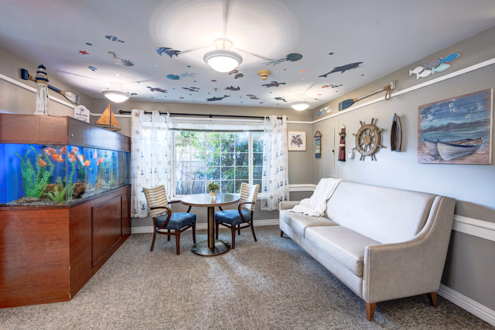 A cozy living room with a large fish tank on the left, a round table with two chairs in front of a window with sheer curtains, and a beige sofa with a white throw blanket on the right. The ceiling is decorated with colorful sea creature decals, and the walls feature nautical-themed decorations including a ship's wheel, oars, and paintings of boats and sea life.