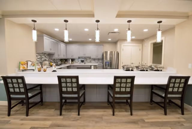 A modern kitchen area with a long white countertop and four wooden chairs with patterned cushions. The kitchen features gray cabinets, a stainless steel refrigerator, a tiled backsplash, and four pendant lights hanging from the ceiling. There are also serving trays on the counter to the right.