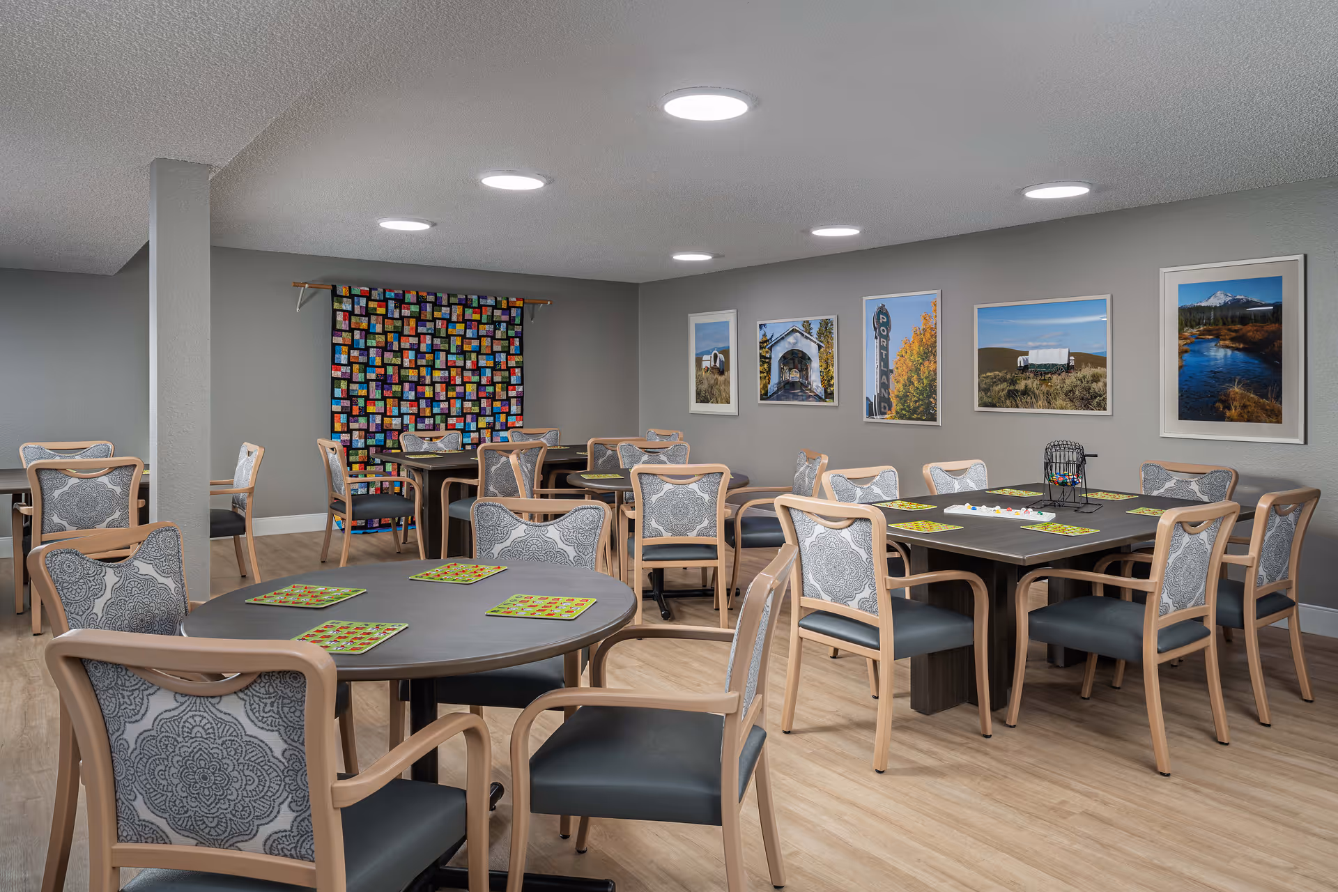 A well-lit room with multiple tables and chairs arranged for group activities or dining. The tables have colorful placemats and a bingo cage with bingo cards on one table. The walls are decorated with framed landscape photographs and a colorful quilt hanging on one wall. The floor is light wood, and the ceiling has several round recessed lights.