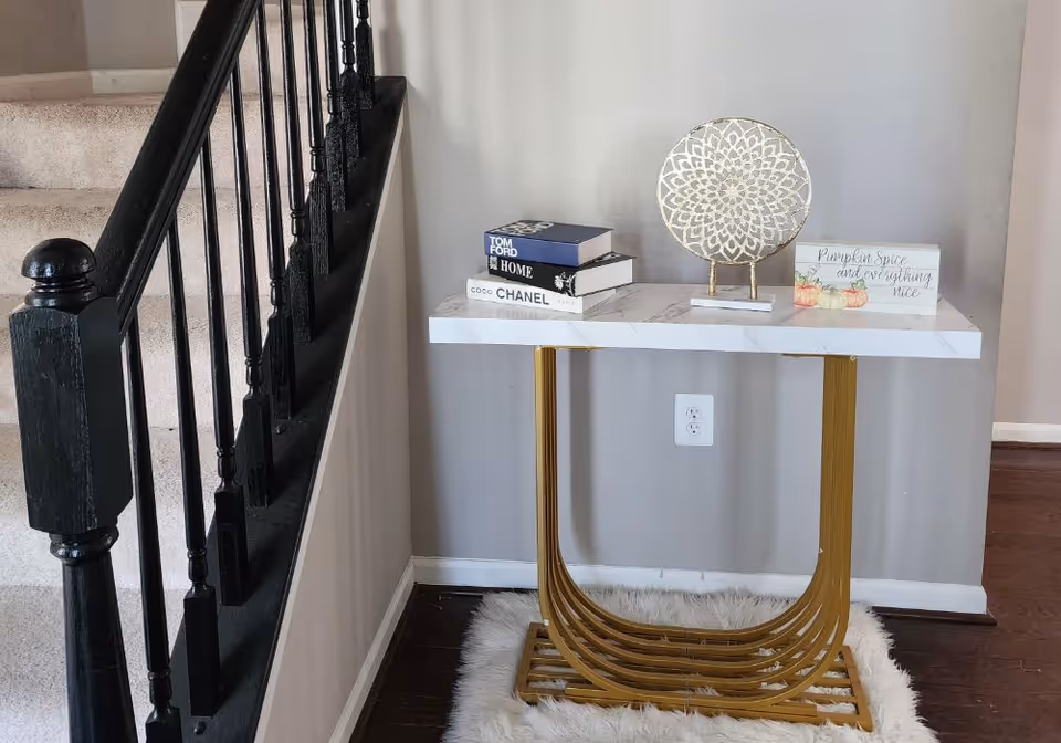 Entryway interior with a black staircase railing next to a marble-top gold console table topped with books and decorative accents.