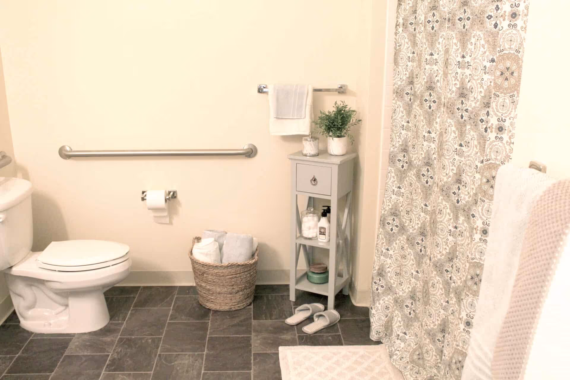 A clean bathroom with a white toilet, a silver grab bar on the wall, a toilet paper holder with a roll of toilet paper, a woven basket with folded towels, a small gray cabinet holding toiletries and a potted plant, a pair of white slippers on the floor, a patterned shower curtain, and a white bath mat on dark tiled flooring.