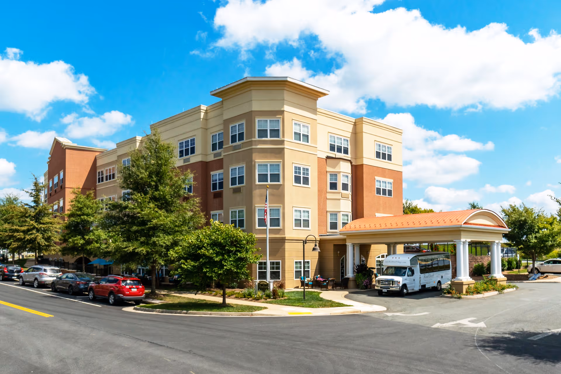 Exterior view of a multi-story assisted living facility building with a covered entrance and a shuttle bus parked in front. Several cars are parked along the street, and trees and landscaping surround the building under a partly cloudy blue sky.