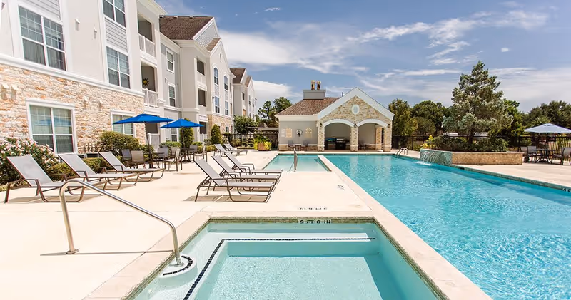Outdoor swimming pool area at The Grand Parkway facility with lounge chairs, umbrellas, a hot tub, and a small building with arches in the background under a partly cloudy sky.