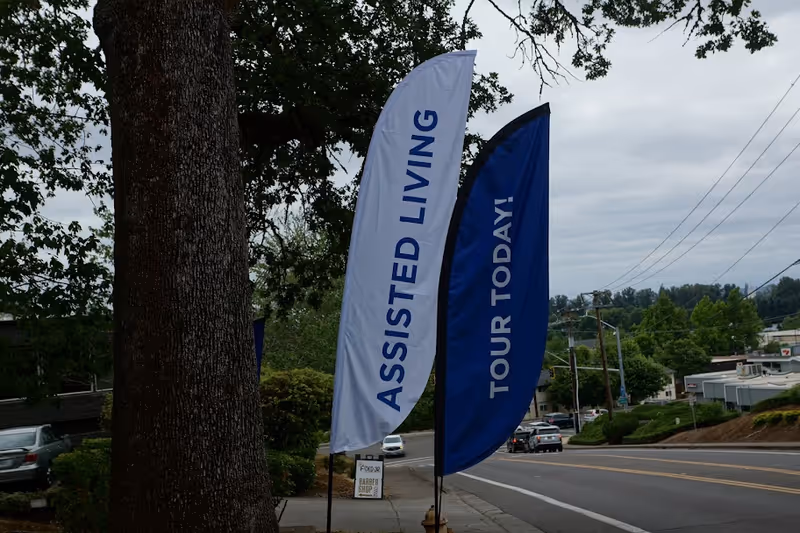 Two feather flags reading 'ASSISTED LIVING' and 'TOUR TODAY!' on a sidewalk beside a street with trees and cars.