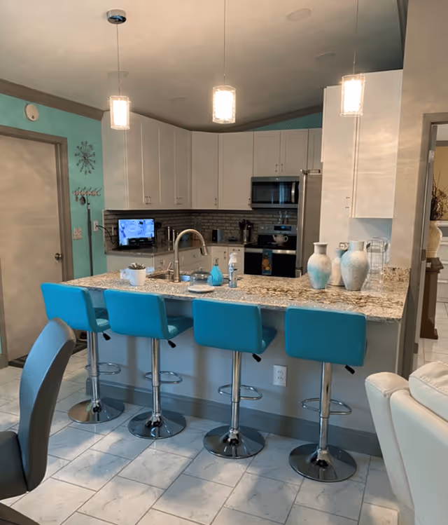 Modern kitchen with a granite countertop island featuring four turquoise bar stools. The kitchen has white cabinets, a stainless steel microwave, and a refrigerator. Three pendant lights hang above the island. Decorative vases and small plants are placed on the countertop. The walls are painted in a light turquoise color with a door on the left side.