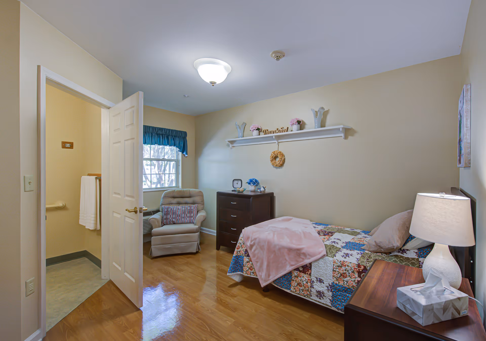 A cozy bedroom in a senior living facility with a single bed covered in a colorful patchwork quilt and a pink blanket. There is a wooden nightstand with a white lamp and a tissue box. A dark wooden dresser stands against the wall, decorated with small flower pots and a clock. A comfortable beige armchair with a patterned pillow is placed near a window with a blue valance. The door to an adjacent bathroom is open, showing a towel hanging on a rail. The walls are painted light beige, and the floor is polished wood.