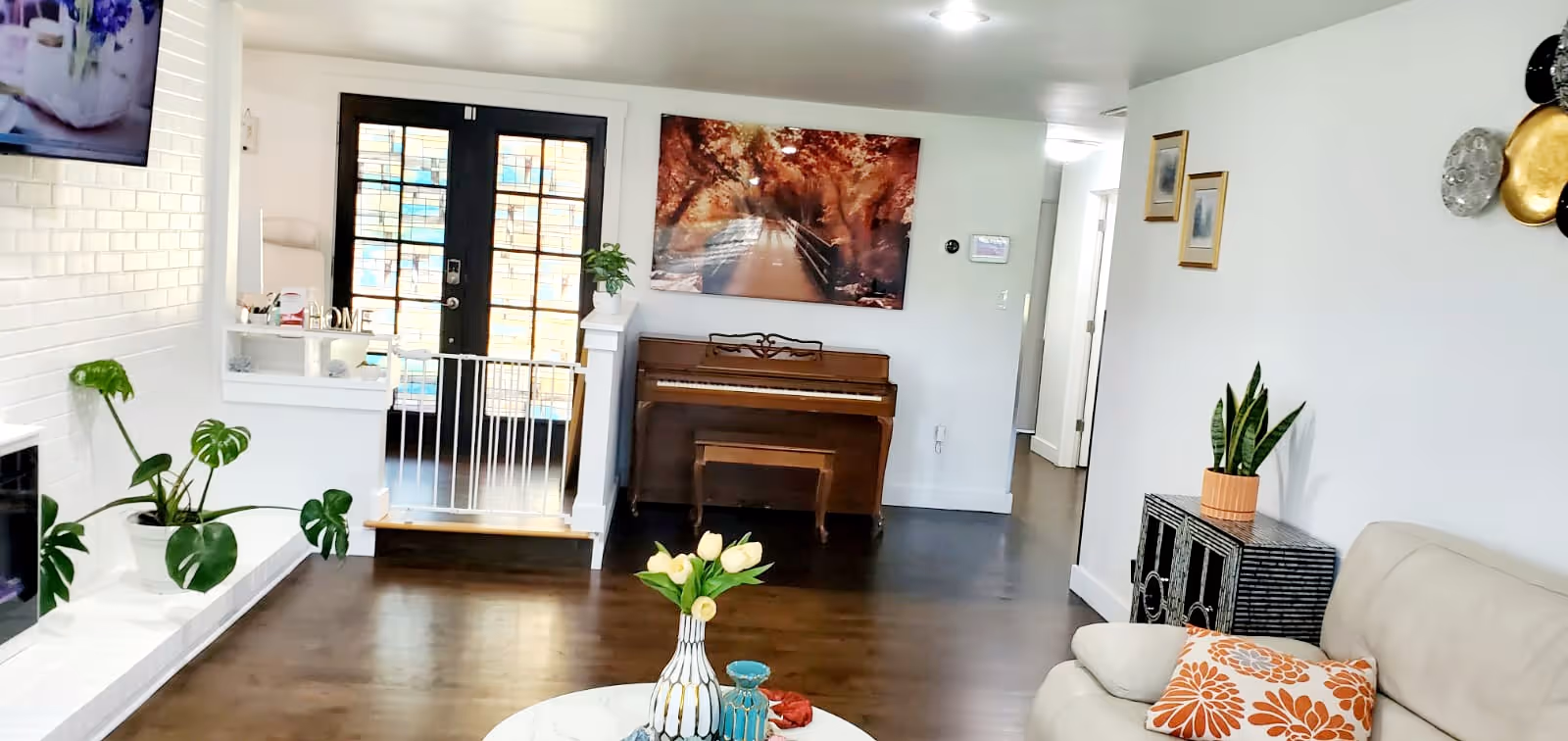 A bright and cozy living room with a white couch adorned with an orange floral pillow, a small black cabinet with a potted plant, and a wooden piano against the far wall. Above the piano is a large framed picture of a tree-lined path with autumn foliage. The room has dark wooden floors and white walls. A white baby gate is positioned in front of a door with glass panels, and a white brick fireplace with green plants on the hearth is visible on the left side. A round white marble coffee table with decorative vases and flowers is in the foreground.
